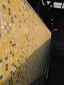 A close-up view of a wall covered with small square tiles arranged in a mosaic pattern. The tiles feature various shades of yellow, green, and gray, and sunlight casts a shadow along the wall. The setting appears to be indoors with some visible railing indicating stairs or a walkway.
