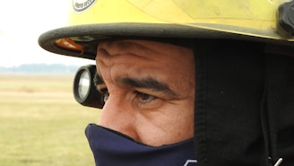 A close-up view of a person wearing a yellow firefighter helmet with a flashlight attached and a protective mask covering the lower half of the face. The background is a blurred outdoor landscape, indicating the person is possibly in a field.