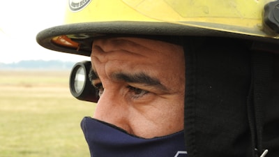 A close-up view of a person wearing a yellow firefighter helmet with a flashlight attached and a protective mask covering the lower half of the face. The background is a blurred outdoor landscape, indicating the person is possibly in a field.