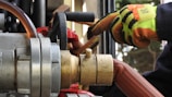 Close-up of industrial equipment showing a metallic valve connected to a thick hose. A person wearing bright orange and yellow protective gloves is operating the machinery. Background includes various mechanical components and structural elements.