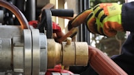 Close-up of industrial equipment showing a metallic valve connected to a thick hose. A person wearing bright orange and yellow protective gloves is operating the machinery. Background includes various mechanical components and structural elements.
