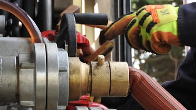 Close-up of industrial equipment showing a metallic valve connected to a thick hose. A person wearing bright orange and yellow protective gloves is operating the machinery. Background includes various mechanical components and structural elements.
