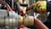 Close-up of industrial equipment showing a metallic valve connected to a thick hose. A person wearing bright orange and yellow protective gloves is operating the machinery. Background includes various mechanical components and structural elements.