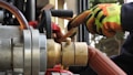 Close-up of industrial equipment showing a metallic valve connected to a thick hose. A person wearing bright orange and yellow protective gloves is operating the machinery. Background includes various mechanical components and structural elements.