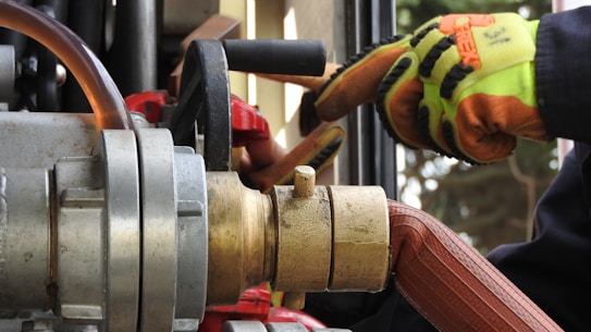 Close-up of industrial equipment showing a metallic valve connected to a thick hose. A person wearing bright orange and yellow protective gloves is operating the machinery. Background includes various mechanical components and structural elements.