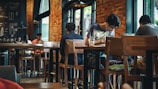 A cozy coffee shop interior with several people sitting at wooden tables. Brick walls and large windows create a warm atmosphere. Some patrons are focused on laptops or books, while others are engaged in conversation. The space is softly lit with hanging lights and has a relaxed vibe.