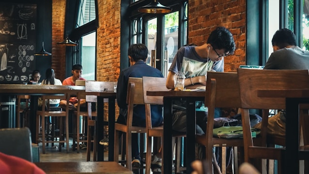 A cozy coffee shop interior with several people sitting at wooden tables. Brick walls and large windows create a warm atmosphere. Some patrons are focused on laptops or books, while others are engaged in conversation. The space is softly lit with hanging lights and has a relaxed vibe.