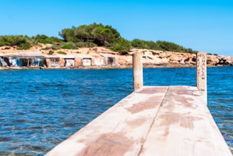 A wooden pier extends over clear blue water with a sign indicating shallow depth in both Spanish and English. In the background, there is a rocky shoreline with small structures and green shrubs.