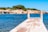 A wooden pier extends over clear blue water with a sign indicating shallow depth in both Spanish and English. In the background, there is a rocky shoreline with small structures and green shrubs.