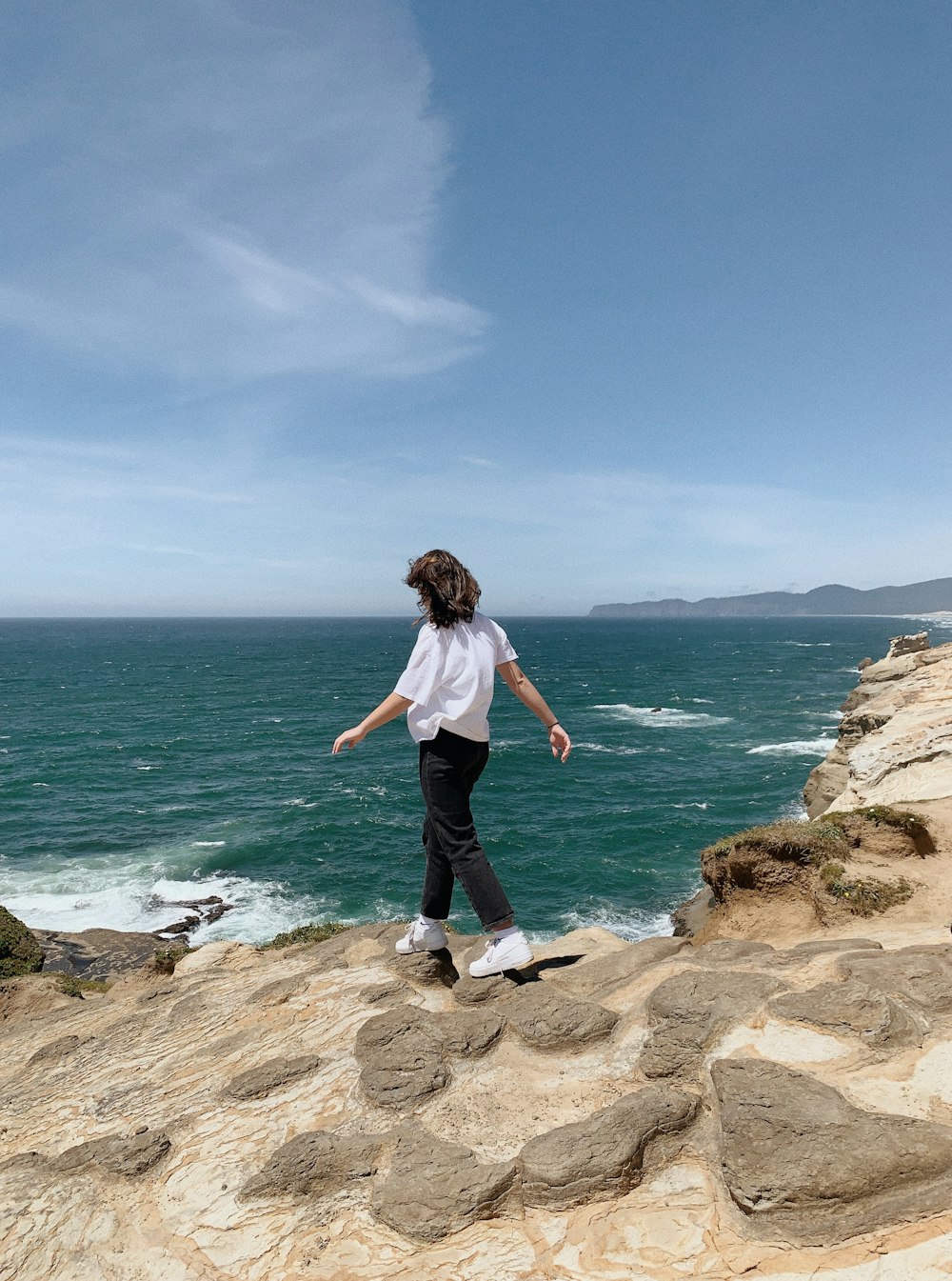 woman in white shirt and black pants standing on brown rock formation near body of water