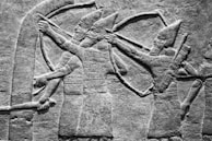 An ancient stone relief depicting two armed figures drawing bows, wearing detailed armor and pointed helmets, portrayed in a profile view typical of ancient artworks.