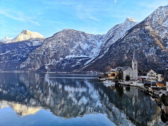 A picturesque lakeside village with a tall church spire nestled against majestic snow-covered mountains. The tranquil lake perfectly mirrors the stunning landscape and clear blue sky.