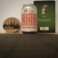 A can labeled 'Popcorn Pimp' with a design featuring red and white stripes and a logo depicting a pile of popcorn on top, alongside a cork coaster with a silhouette of a city labeled 'Riga'. In the background, there is a book with an illustration of a fictional creature on a green cover, showing text and some small stars.