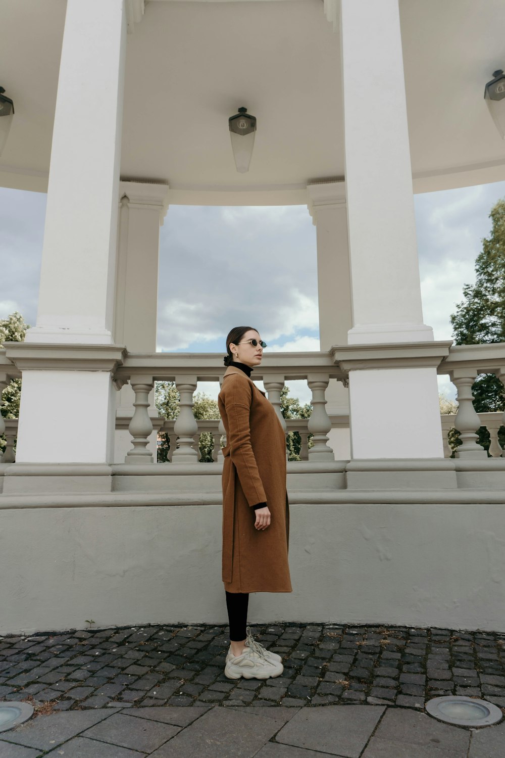 woman in brown coat standing near white concrete building during daytime