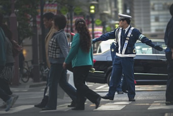 A police officer in uniform is directing traffic on a busy city street. Several pedestrians are crossing the street, and a black car is visible in the background. The scene is bustling and urban, with various people moving in different directions.