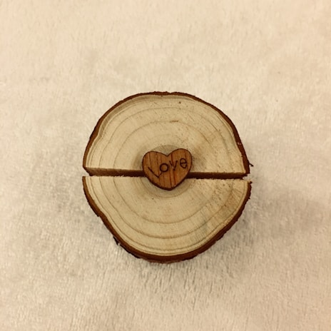 A wooden slice with concentric rings is placed on a light fabric background. At the center, a small heart-shaped piece with the word 'Love' engraved on it is embedded in the wood.