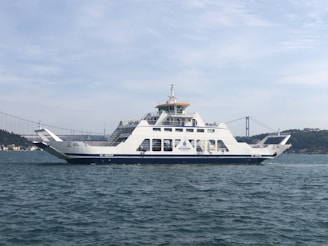 A large ferry is sailing on a body of water, with a suspension bridge and a distant city skyline in the background. The ferry is white with blue accents and appears to be carrying passengers. The water is slightly wavy, and the sky is mostly clear with a few clouds.