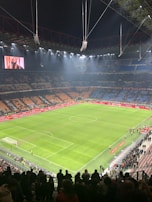 An illuminated football stadium with an expansive, well-maintained grass pitch surrounded by spectator stands. The image shows a large scoreboard with a digital screen displaying an image. Few people are on the field, while more spectators gather near the edges or in the stands.