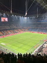 An illuminated football stadium with an expansive, well-maintained grass pitch surrounded by spectator stands. The image shows a large scoreboard with a digital screen displaying an image. Few people are on the field, while more spectators gather near the edges or in the stands.