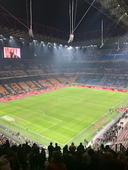 An illuminated football stadium with an expansive, well-maintained grass pitch surrounded by spectator stands. The image shows a large scoreboard with a digital screen displaying an image. Few people are on the field, while more spectators gather near the edges or in the stands.