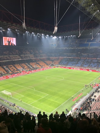 An illuminated football stadium with an expansive, well-maintained grass pitch surrounded by spectator stands. The image shows a large scoreboard with a digital screen displaying an image. Few people are on the field, while more spectators gather near the edges or in the stands.