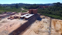 A construction site features multiple structures in various stages of development, with brick buildings prominently situated in an area cleared of vegetation. Workers are visible on-site carrying out construction tasks. The site is surrounded by greenery and trees, with hills and other built structures in the distance.