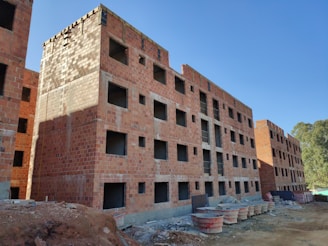 A construction site featuring multiple unfinished brick apartment buildings. The buildings have numerous square window openings and lack windows or doors. The site is surrounded by construction materials and debris. The sky is clear and blue with visible trees on the right side.
