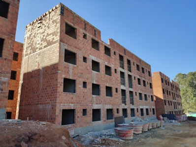 A construction site featuring multiple unfinished brick apartment buildings. The buildings have numerous square window openings and lack windows or doors. The site is surrounded by construction materials and debris. The sky is clear and blue with visible trees on the right side.