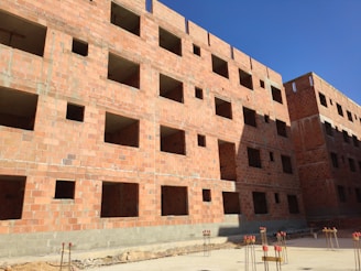 A multi-story building under construction with red brick walls. The structure features numerous window openings and lacks window installations. The building is surrounded by construction materials, including metal rods and concrete. The sky is clear and blue, suggesting a sunny day.