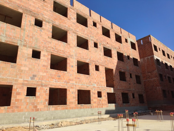 A multi-story building under construction with red brick walls. The structure features numerous window openings and lacks window installations. The building is surrounded by construction materials, including metal rods and concrete. The sky is clear and blue, suggesting a sunny day.