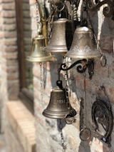 A collection of vintage bells of varying sizes and designs hanging on a textured brick wall. The bells are metallic, featuring engraved designs and numbers like '1824'. Some are attached with chains, and there's a framed decorative piece on the wall next to them.