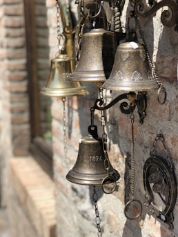 A collection of vintage bells of varying sizes and designs hanging on a textured brick wall. The bells are metallic, featuring engraved designs and numbers like '1824'. Some are attached with chains, and there's a framed decorative piece on the wall next to them.