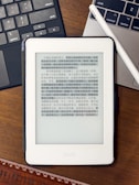 A Kindle device displaying text in Chinese is placed on a wooden table. Nearby, a laptop keyboard on the left and partially visible on the right is a white stylus resting on the laptop. At the bottom of the image, part of a wooden ruler is visible.