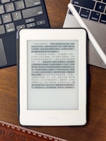 A Kindle device displaying text in Chinese is placed on a wooden table. Nearby, a laptop keyboard on the left and partially visible on the right is a white stylus resting on the laptop. At the bottom of the image, part of a wooden ruler is visible.
