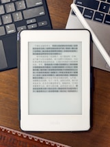A Kindle device displaying text in Chinese is placed on a wooden table. Nearby, a laptop keyboard on the left and partially visible on the right is a white stylus resting on the laptop. At the bottom of the image, part of a wooden ruler is visible.