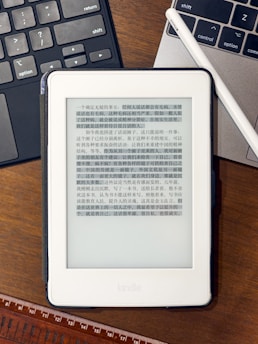 A Kindle device displaying text in Chinese is placed on a wooden table. Nearby, a laptop keyboard on the left and partially visible on the right is a white stylus resting on the laptop. At the bottom of the image, part of a wooden ruler is visible.
