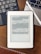 A Kindle device displaying text in Chinese is placed on a wooden table. Nearby, a laptop keyboard on the left and partially visible on the right is a white stylus resting on the laptop. At the bottom of the image, part of a wooden ruler is visible.