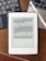 A Kindle device displaying text in Chinese is placed on a wooden table. Nearby, a laptop keyboard on the left and partially visible on the right is a white stylus resting on the laptop. At the bottom of the image, part of a wooden ruler is visible.