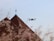 A drone flies through the air near a rooftop with a weather vane, surrounded by soft-focus pink blossoms. The sky is clear blue, providing a serene backdrop.
