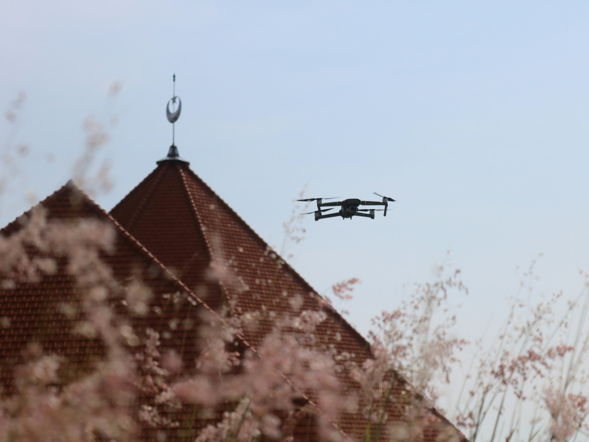 A drone flies through the air near a rooftop with a weather vane, surrounded by soft-focus pink blossoms. The sky is clear blue, providing a serene backdrop.
