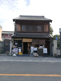 A traditional Japanese building with a wooden facade and tiled roof. The ground floor has an open entrance, and several signs and posters are displayed outside. Ice cream cone replicas and other decorative items are present at the entrance. The building is located on a quiet street with some greenery visible to the right.