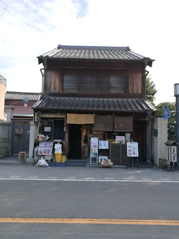 A traditional Japanese building with a wooden facade and tiled roof. The ground floor has an open entrance, and several signs and posters are displayed outside. Ice cream cone replicas and other decorative items are present at the entrance. The building is located on a quiet street with some greenery visible to the right.