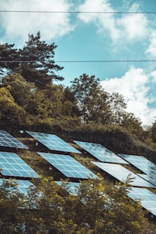 Solar panels are installed on a hillside surrounded by lush green trees under a partly cloudy sky.