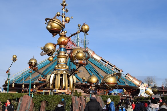A futuristic, steampunk-inspired structure with large, metallic orbs and spires sits prominently. The design includes intricate machinery and clockwork elements, with a pointed roof adorned with vibrant colors and patterns. People are gathered around, engaging in leisurely activities.