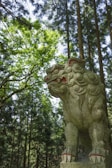 A stone statue of a mythical creature resembling a lion with intricate carvings and red accents stands in a forest. The statue is surrounded by tall trees, with sunlight filtering through the lush green foliage, casting dappled light and shadow on the sculpture.