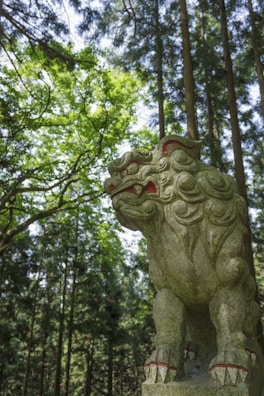 A stone statue of a mythical creature resembling a lion with intricate carvings and red accents stands in a forest. The statue is surrounded by tall trees, with sunlight filtering through the lush green foliage, casting dappled light and shadow on the sculpture.
