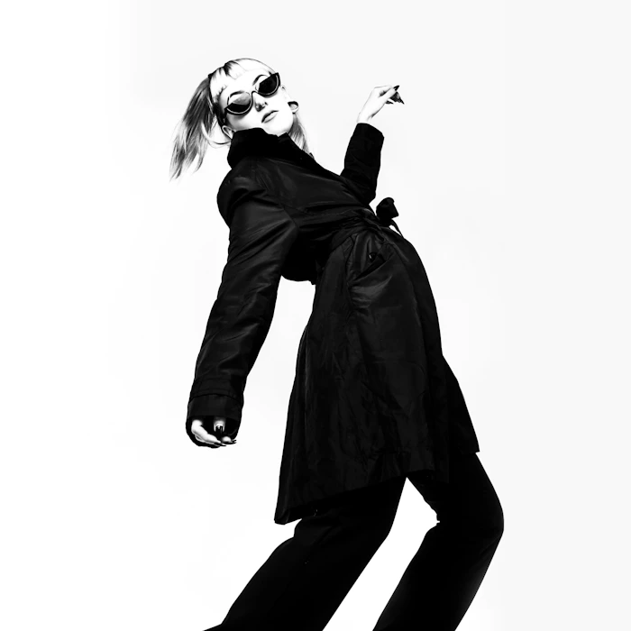 Model in a long black coat leaning mid-stride against a white backdrop