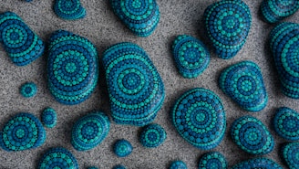 A collection of stones of varying sizes is laid out on a textured surface. Each stone is intricately painted with concentric circular patterns using shades of blue and teal, creating a visually appealing mosaic effect.