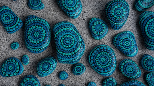 A collection of stones of varying sizes is laid out on a textured surface. Each stone is intricately painted with concentric circular patterns using shades of blue and teal, creating a visually appealing mosaic effect.
