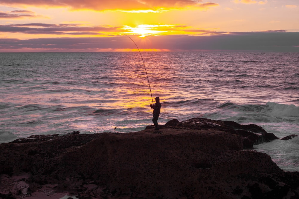 silhouette of person holding fishing rod standing on rock near sea during sunset